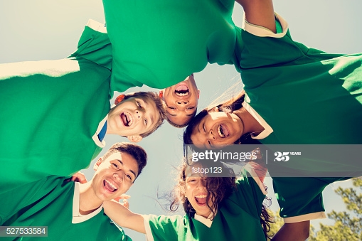 Multi-ethnic team of teenage friends, soccer sports players with park, soccer field and sky  in background.  The team wear green uniforms and huddle together.  Low angle view.  The children cheer together after winning their game.  Go team!   Spring or summer season.